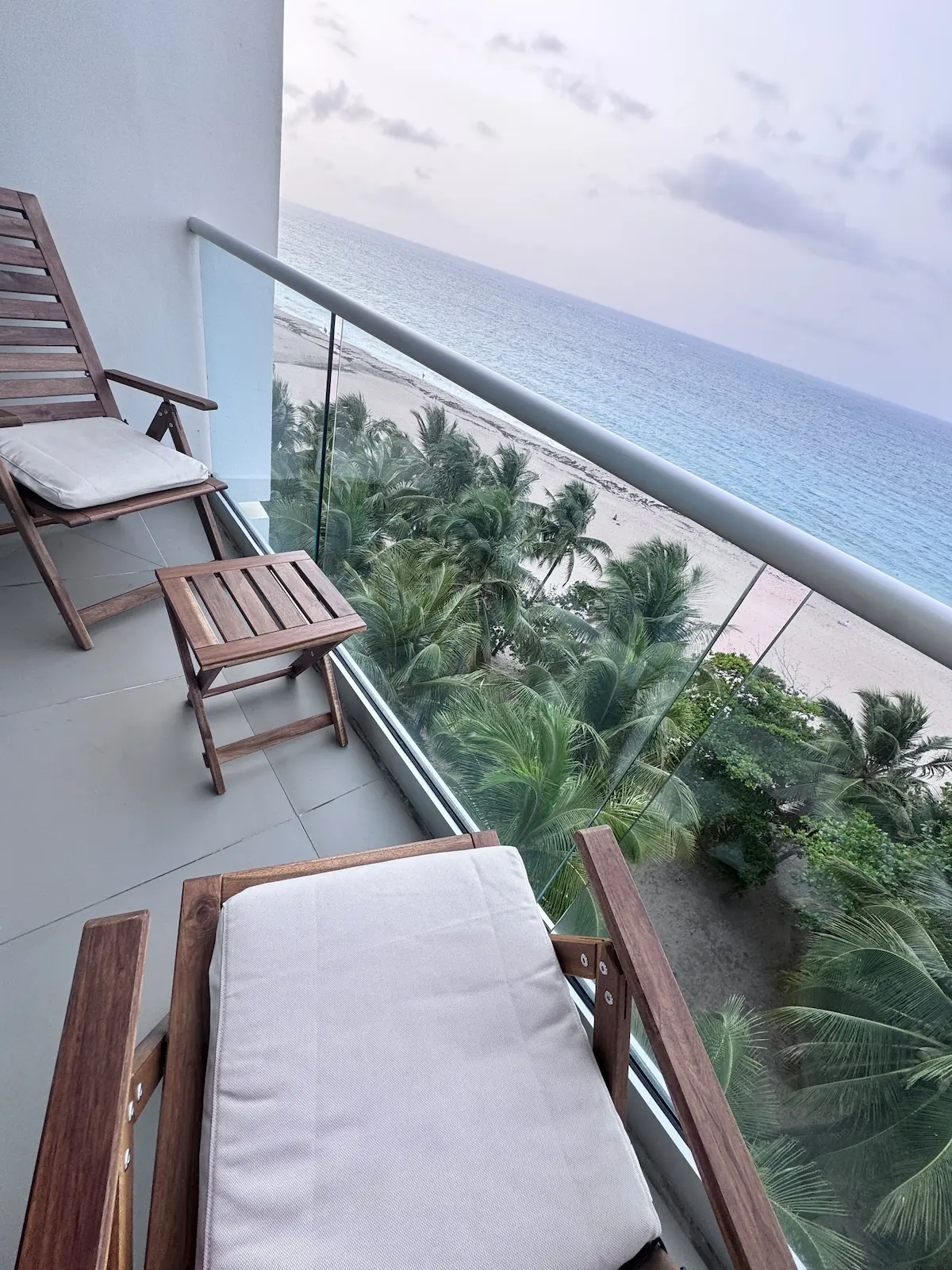 High-floor balcony with two wooden lounge chairs and a glass railing, palm trees below, and the ocean and beach beyond.