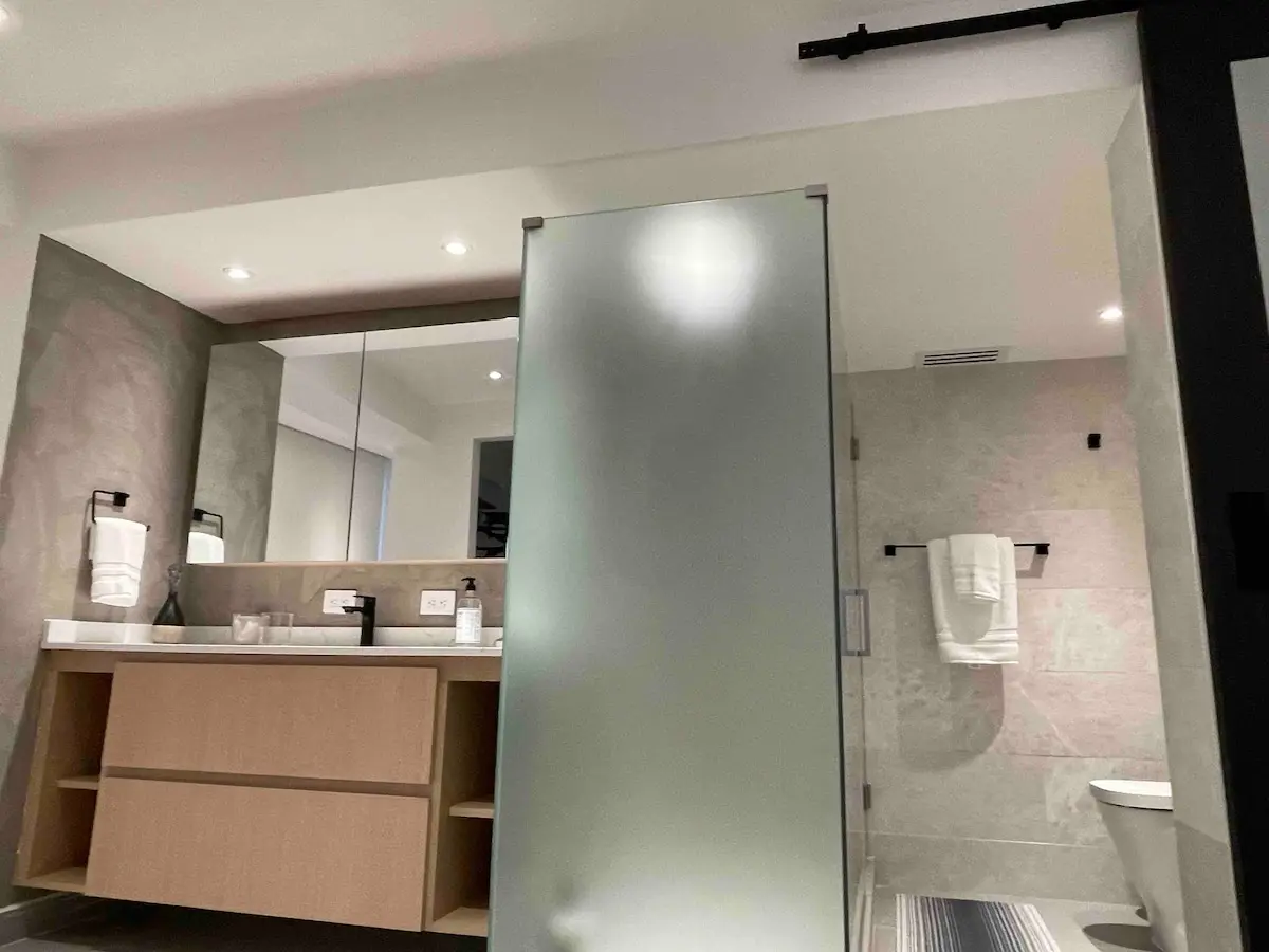 The other full bathroom, with a wood vanity, a white countertop, a large wall mirror, folded towels on a bar, and a frosted glass sliding door onto the shower.