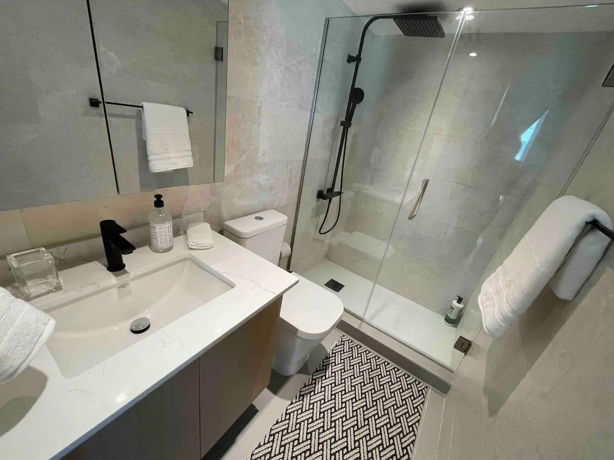 Full bathroom with a white vanity and integrated sink, a toilet, a glass walk-in shower with a black rainfall head, and a black and white patterned tile floor.