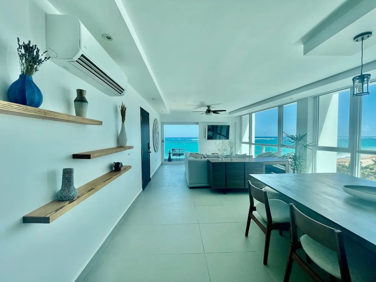Dining table beside floating wood shelves, with floor to ceiling windows looking out to turquoise water.