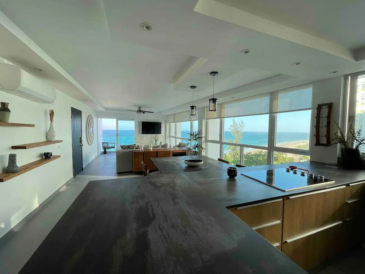 Open-plan kitchen with a dark stone island and pendant lights, and floor to ceiling windows facing the ocean.