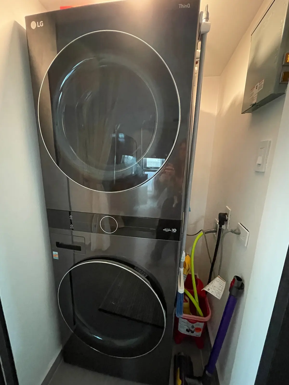 In-unit laundry closet with a stacked dark grey front-load washer and dryer, cleaning tools hanging alongside, and a wall control panel.
