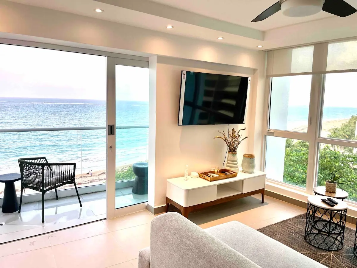 Living room with a wall-mounted TV and ceiling fan, the sliding glass door open to the balcony and the ocean beyond.