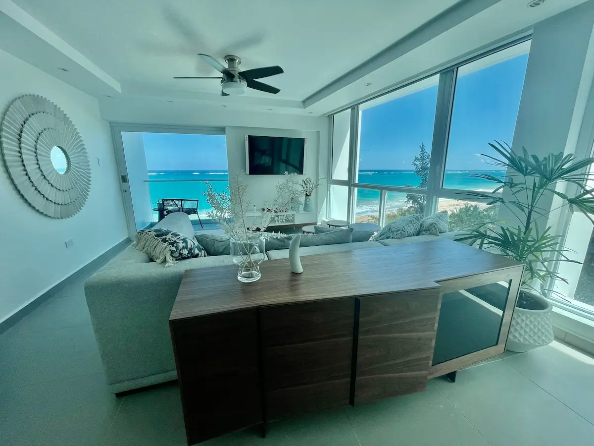 Living room with a light sectional sofa, a wall-mounted TV and a wood coffee table, a ceiling fan overhead, and floor to ceiling windows with the balcony door open to the ocean.