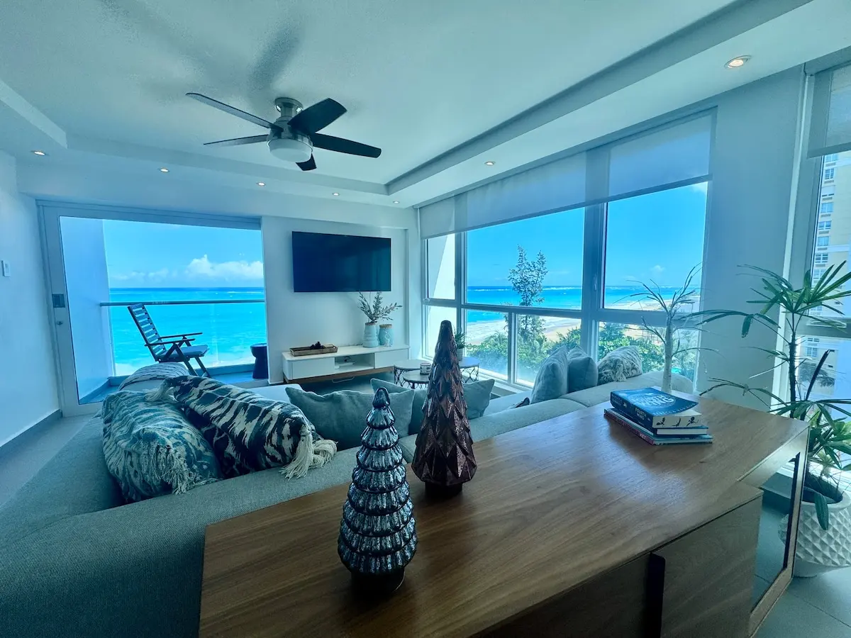 Living room with a light sectional sofa, a wall-mounted TV, a wood coffee table and a ceiling fan, the sliding door open to a balcony with a lounge chair on it.
