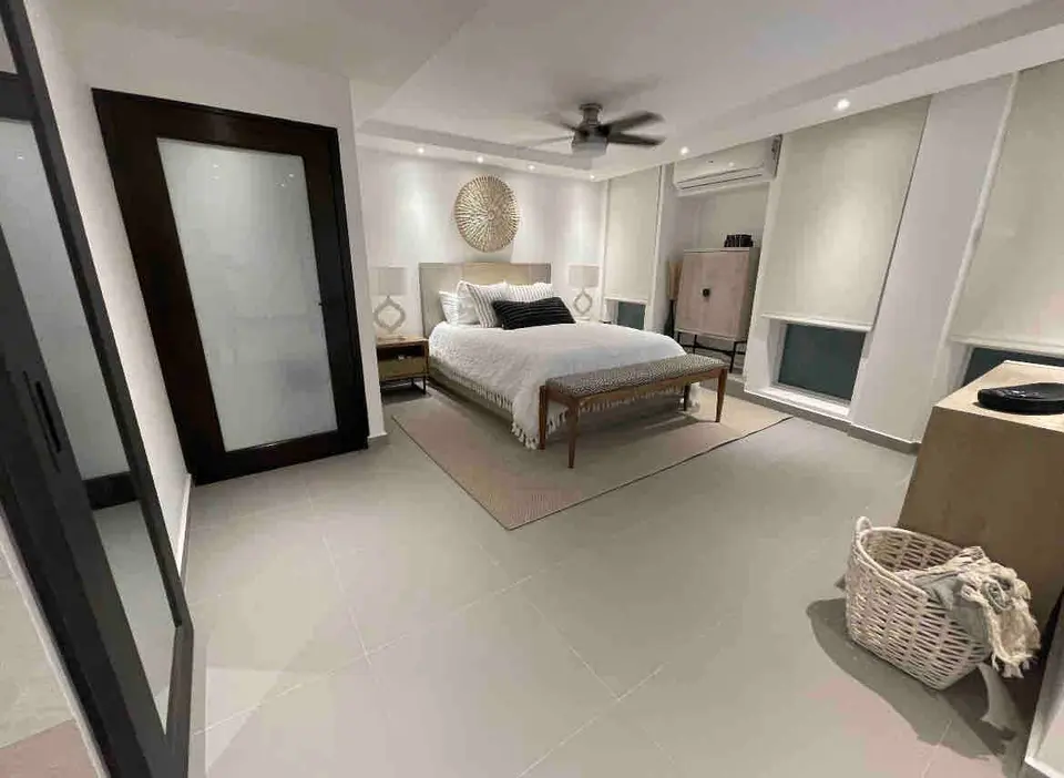 Master bedroom with a white-dressed bed and a bench at the foot, twin nightstands with lamps, a sunburst mirror above the headboard, and a ceiling fan.