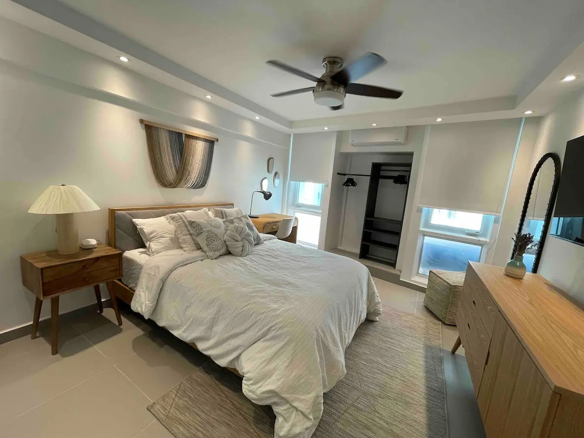 Second bedroom with a white-dressed bed, a wooden nightstand and lamp, a dresser, a wall-mounted TV, and a window with sheer curtains onto turquoise water.